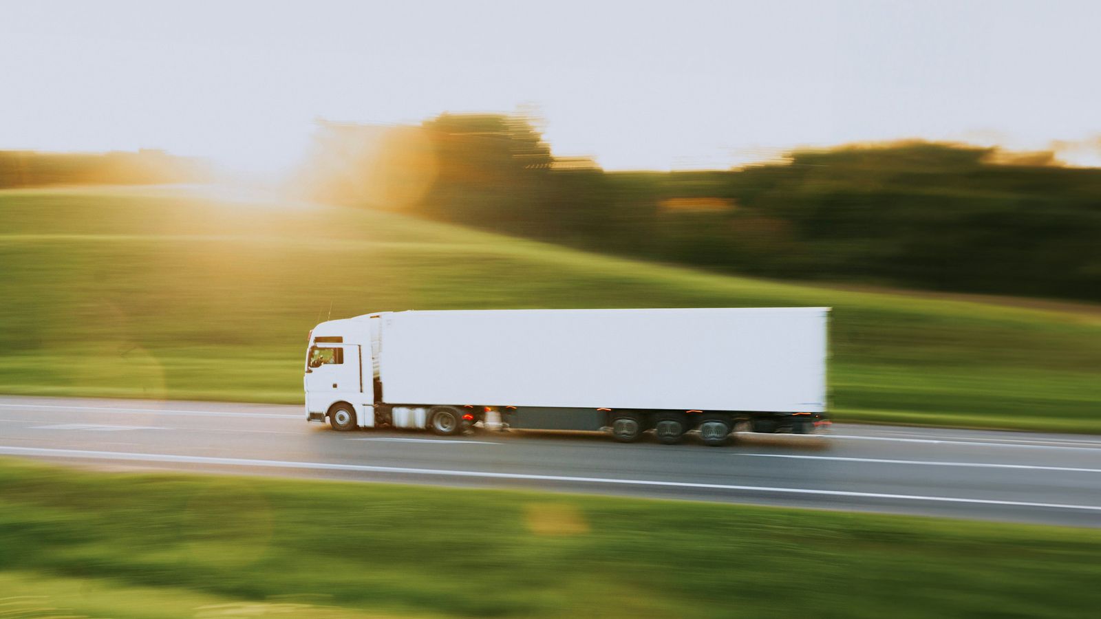Een witte vrachtwagen met een witte oplegger rijdt over een weg tussen groene velden, de achtergrond vervaagt door de snelheid